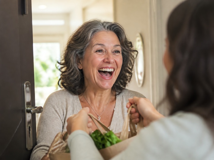 A woman in her fifties laughing at her doorway as a friend takes the heavy bags from her