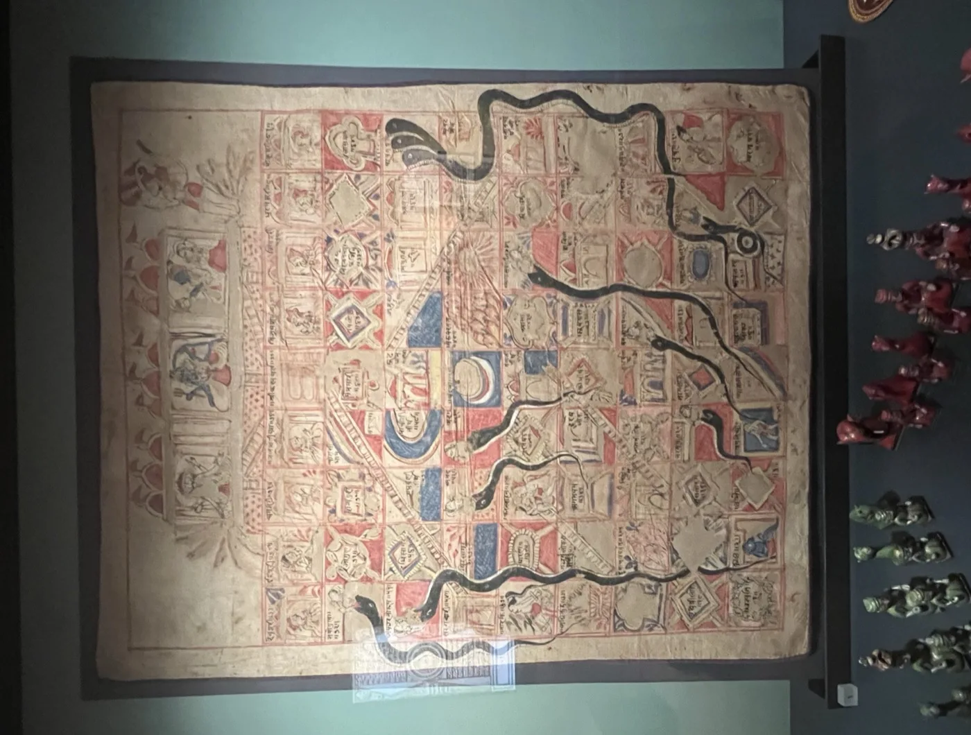 A large cloth gyan chaupar chart mounted on a museum wall, with deities painted across the top row and black snakes winding down the grid