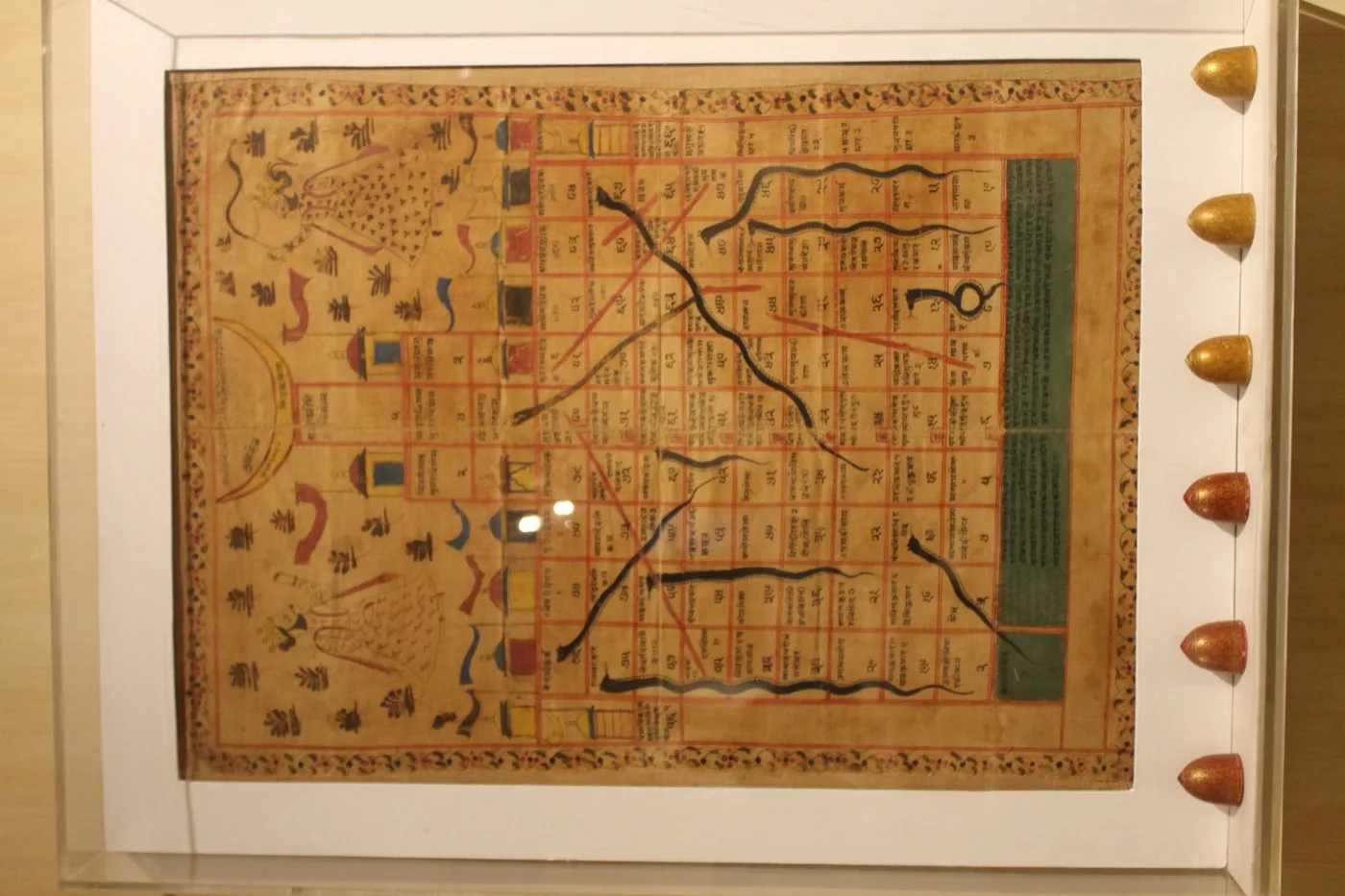 An 84-square Jain gyan chaupar chart in a display case, numbered squares in Devanagari with black snakes and red ladders